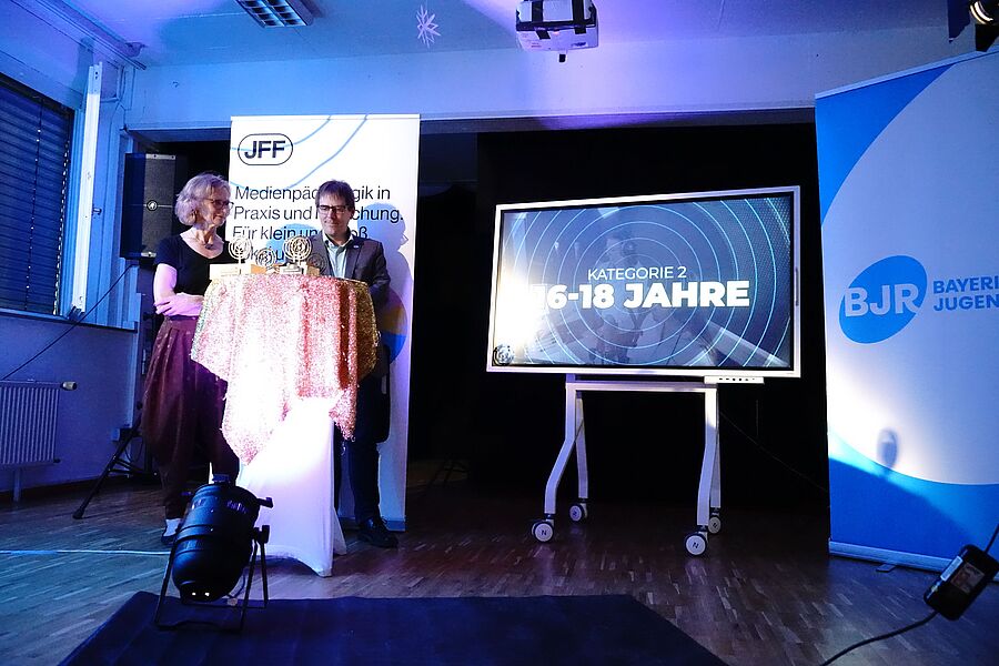 Zwei Personen stehen an einem Tisch mit goldenem Tuch, lächeln und halten einen Preis. Im Hintergrund ist ein Banner mit dem Logo des JFF zu sehen.